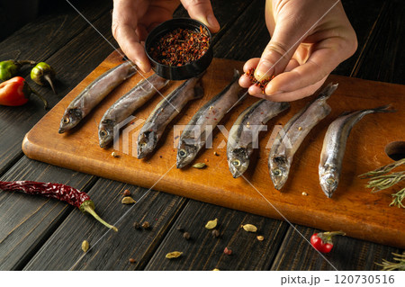 Hands skillfully sprinkle spices over fresh fish lined on a wooden board, surrounded by vibrant herbs and chilies. A fusion of flavors awaits in this culinary creation. Hands skillfully sprinkle spices over fresh fish lined on a wooden board, surrounded by vibrant herbs and chilies. A fusion of flavors awaits in this culinary creation. 120730516