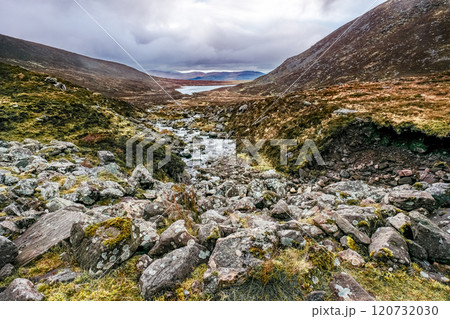 A stream flowing through a rocky valley. The stream is surrounded by rocks, covered in moss and grass. The valley is narrow and steep, with hills rising up on either side. The sky is overcast. A stream flowing through a rocky valley. The stream is surrounded by rocks, covered in moss and grass. The valley is narrow and steep, with hills rising up on either side. The sky is overcast. 120732030