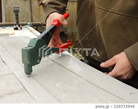 A worker fastens the gray flat metal plates lying on the table with a green riveting tool with red handles 120733044