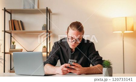 Career man. Technology help. Successful young handsome guy in glasses in black shirt holding smartphone before laptop sitting in office online using gadget. 120733141