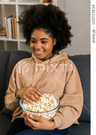 African american woman sits on a gray sofa, enjoying a bowl of popcorn while watching a movie or streaming online film. Cozy and relaxing moment of entertainment in a modern home. Connection with 120733634