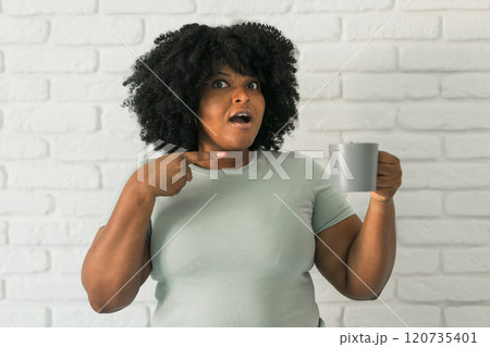 African American surprised woman holding a gray mug while pointing at herself, conveying emotions of shock and curiosity. Moments of self-realization, humor, and unexpected situations in daily life 120735401