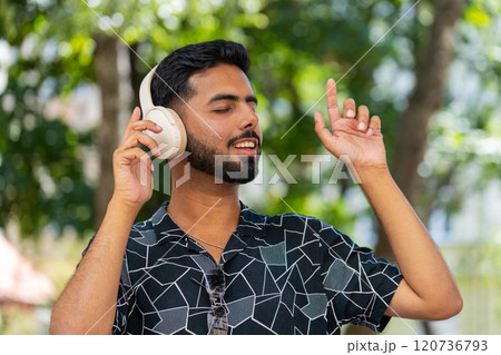 Happy relaxed young Indian man in wireless headphones listening disco music dancing outdoors Happy relaxed young Indian man in wireless headphones listening disco music dancing outdoors 120736793