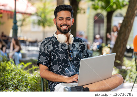 Indian man in headphones working online distant job with laptop browsing website chatting outdoors 120736795