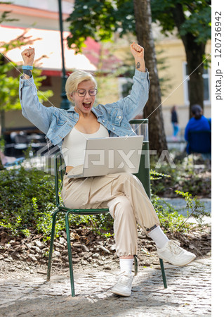 Overjoyed happy young woman working on laptop screaming in delight raises hands in city street 120736942