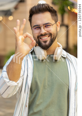 Portrait of handsome young millennial man looking at camera showing hand ok sign on city street Portrait of handsome young millennial man looking at camera showing hand ok sign on city street 120736944
