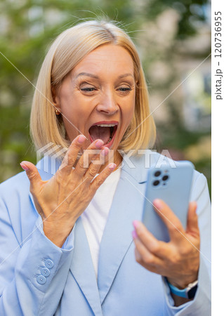 Caucasian mature businesswoman looking shocked while reading good news on smartphone on city street Caucasian mature businesswoman looking shocked while reading good news on smartphone on city street 120736955