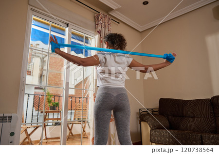 Woman exercising with resistance band at home by large window 120738564