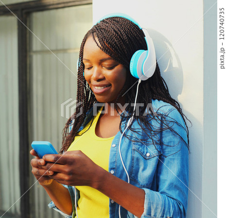 Fashion pretty smiling african woman with headphones listens to music using smartphone in the city 120740735