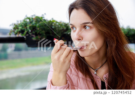 Woman sits savoring her meal with spoon she has long hair is dressed warmly green plants are visible in creating cozy atmosphere. Woman sits savoring her meal with spoon she has long hair is dressed warmly green plants are visible in creating cozy atmosphere. 120745452