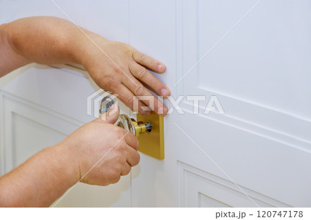 Carpenter is carefully installing door lock on white panel door, during home improvement project. 120747178