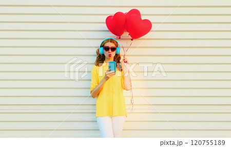 Happy young woman with smartphone, red heart balloons listening to music in headphones on white 120755189