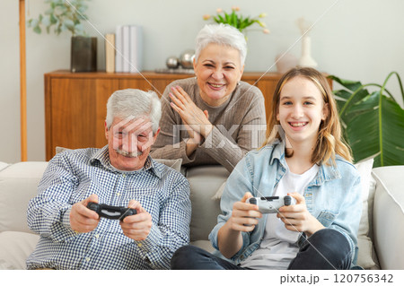 Happy family at home. Old senior man grandfather woman grandmother girl granddaughter playing video game with joysticks at home. Grandparent teenager two generations using gamepads for video game Happy family at home. Old senior man grandfather woman grandmother girl granddaughter playing video game with joysticks at home. Grandparent teenager two generations using gamepads for video game 120756342