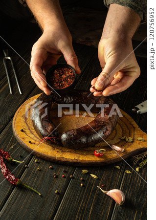 Hands carefully sprinkle spices onto a juicy dark blood sausage lying on a wooden board, surrounded by garlic and herbs, demonstrating the art of traditional cooking in a cozy kitchen 120756952
