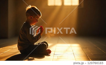 child sits on the floor, hands clasped together in prayer with sunlight streaming 120760183