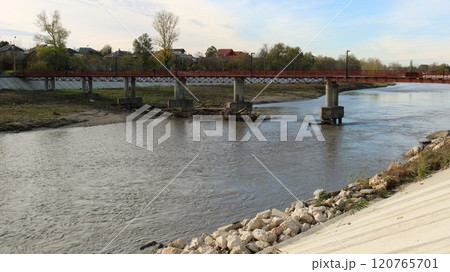 a small red iron bridge over a river with a calm flow of water leading from the bank with a concrete slope and a stone embankment to a residential rural area with trees a small red iron bridge over a river with a calm flow of water leading from the bank with a concrete slope and a stone embankment to a residential rural area with trees 120765701