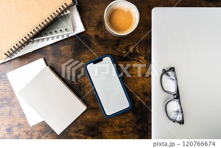 Workspace with smartphone in blue case, spiral notebooks, blank paper, glasses, coffee cup, and laptop on wooden desk 120766674