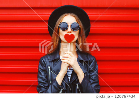 Fashion pretty young woman with red lollipop heart in black hat leather jacket over wall background 120767434