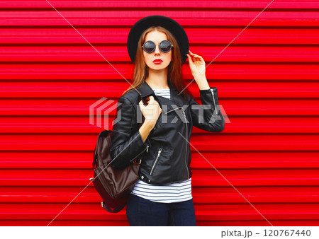 Fashion portrait young woman in black rock style hat, jacket stands with backpack on red background 120767440