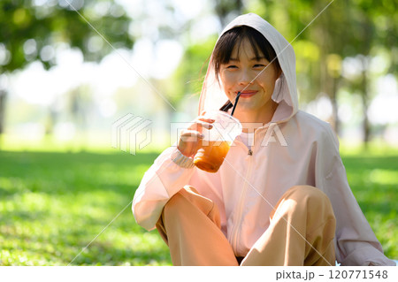 Cheerful young woman sitting on the grass in a park and enjoying refreshing drink after exercising 120771548