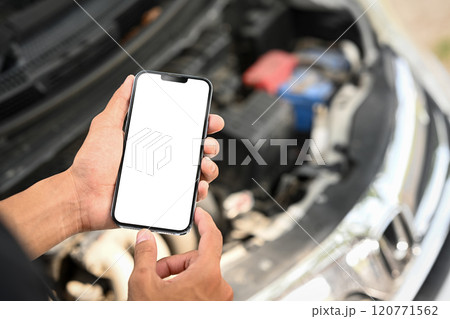 Man using smartphone near an open car hood while inspecting the car engine Man using smartphone near an open car hood while inspecting the car engine 120771562