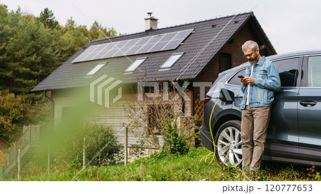 Man charging electric car in front of his house with solar panel system on roof behind him. Man leaning against a car while scrolling on smartphone. Man charging electric car in front of his house with solar panel system on roof behind him. Man leaning against a car while scrolling on smartphone. 120777653