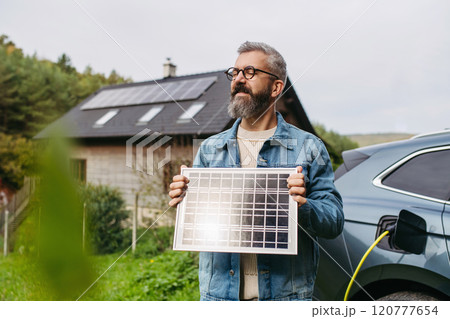 Man holding model of solar panel in hands. Charging electric car and house with solar panel system on roof behind him. 120777654