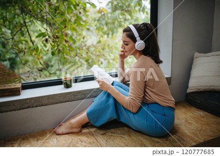 Woman sitting by a large window with headphones on head and book. Woman is enjoying time for herself, reading. Woman sitting by a large window with headphones on head and book. Woman is enjoying time for herself, reading. 120777685