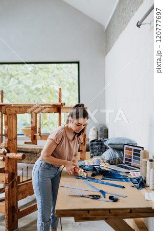 Young businesswoman working on workbench, preparing old fabric for weaving. Upcycled textile, small weaving business and circular economy. Young businesswoman working on workbench, preparing old fabric for weaving. Upcycled textile, small weaving business and circular economy. 120777697