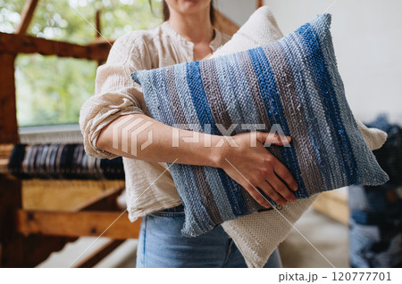 Young businesswoman next to wooden loom, holding pillows with woven covers made from recycled fabrics. Upcycling in business and circular economy. Young businesswoman next to wooden loom, holding pillows with woven covers made from recycled fabrics. Upcycling in business and circular economy. 120777701