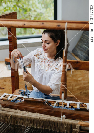 Young businesswoman sitting at a wooden loom, weaving a rug from old fabrics. Upcycling in business and circular economy. Young businesswoman sitting at a wooden loom, weaving a rug from old fabrics. Upcycling in business and circular economy. 120777702
