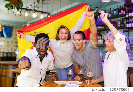 Excited diverse soccer supporters with flag of Spain celebrating victory with pint of beer in the pub Excited diverse soccer supporters with flag of Spain celebrating victory with pint of beer in the pub 120778605