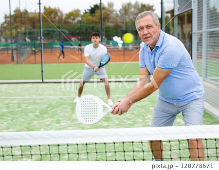 Concentrated elderly woman padel player hitting ball with a racket on hard court 120778671