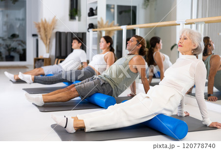 Active senior woman performs exercises with a pilates roller while lying on a mat in the hall of a modern fitness studio. 120778740