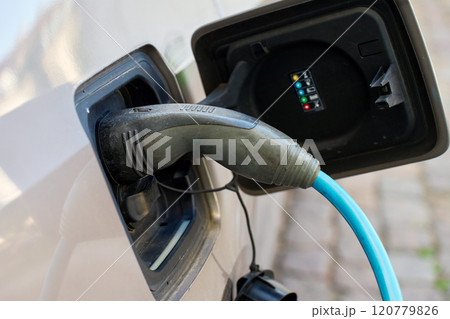 Close up of electric vehicle charging on public station at city street. EV car with plugged in cable recharging battery. Sustainable and eco-friendly transportation Close up of electric vehicle charging on public station at city street. EV car with plugged in cable recharging battery. Sustainable and eco-friendly transportation 120779826