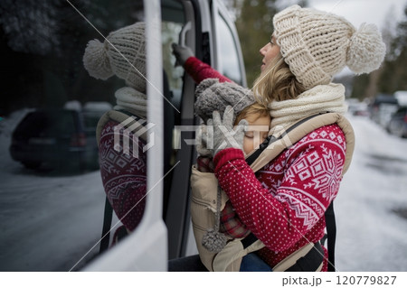 Mother standing in the middle of snowy parking lot with sleeping daughter in baby carrier. Mom and girl enjoying winter holiday in the mountains. 120779827