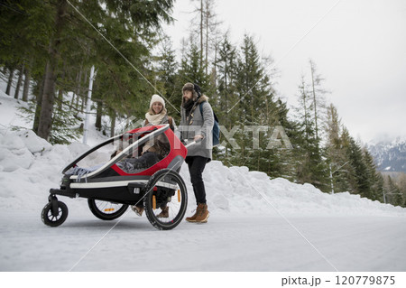 Young family is enjoying winter holiday in the mountains. Parents pushing kids in wagon stroller through snowy nature. 120779875