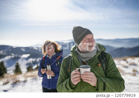 Active seniors resting, drinking warm tea from thermos. Romantic winter hike for an elderly couple in snowy mountains. 120779919