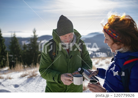 Active seniors resting, drinking warm tea from thermos. Romantic winter hike for an elderly couple in snowy mountains. 120779926