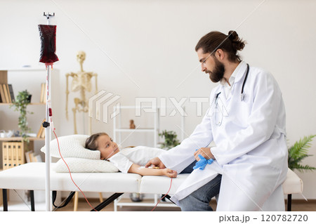 Male doctor provides blood transfusion to young Caucasian girl in medical setting. Child lying on medical bed receiving treatment. Professional healthcare environment, focus on care and comfort. 120782720