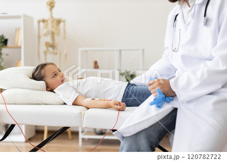 Doctor gives blood transfusion to sick little girl at medical facility. Young girl lying on bed in hospital room receiving treatment from male doctor wearing lab coat. 120782722