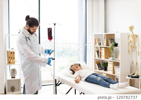 Male doctor provides blood transfusion to Caucasian young girl lying on examination table. Doctor in medical coat and gloves adjusting IV drip in bright hospital room. Male doctor provides blood transfusion to Caucasian young girl lying on examination table. Doctor in medical coat and gloves adjusting IV drip in bright hospital room. 120782737