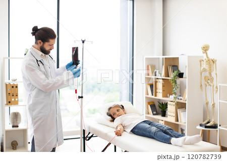 Male doctor provides blood transfusion to young girl of mixed race in clinical setting. Girl lies on examination table, looking calm. Room features medical equipment and skeleton model. Male doctor provides blood transfusion to young girl of mixed race in clinical setting. Girl lies on examination table, looking calm. Room features medical equipment and skeleton model. 120782739