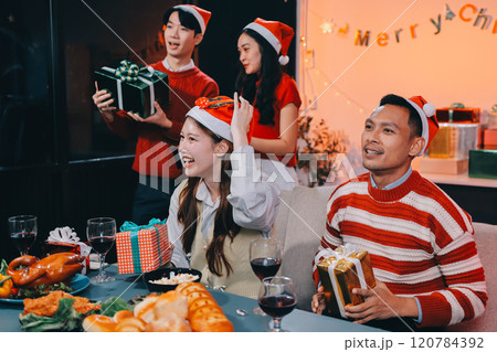Group of happy Asian friends celebrating Christmas and decorate the Christmas tree indoors. Beauty woman with Christmas Gifts. New Year party. Woman hands decorate Christmas tree red ball, bauble. 120784392