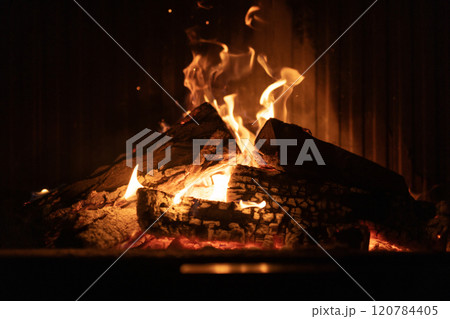 A close-up view of a glowing fireplace, with flames dancing over burning logs. The warm tones and flickering light create a cozy and tranquil ambiance. 120784405