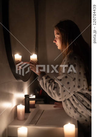 A young woman in soft lighting holds a candle near a mirror, her finger touching her reflection. The warm glow of the candles creates a calm and intimate atmosphere. A young woman in soft lighting holds a candle near a mirror, her finger touching her reflection. The warm glow of the candles creates a calm and intimate atmosphere. 120784439