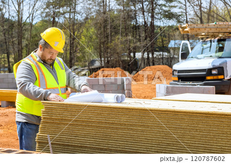 Construction supervisor worker in safety vest, hard hat studies blueprints on stack of materials at residential building site heavy machinery 120787602