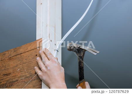 Contractor hand grips hammer while another adjusts electrical wiring against wooden framing in construction area, home improvement efforts. Contractor hand grips hammer while another adjusts electrical wiring against wooden framing in construction area, home improvement efforts. 120787629