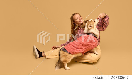 Beautiful young woman in checkered shirt posing with adorable happy Corgi dog wearing pink sweater against warm studio background Beautiful young woman in checkered shirt posing with adorable happy Corgi dog wearing pink sweater against warm studio background 120788607