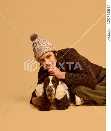 Portrait of young man in warm, cozy chocolate sweater posing with his adorable calm dog wearing warm headwear, against beige studio background Portrait of young man in warm, cozy chocolate sweater posing with his adorable calm dog wearing warm headwear, against beige studio background 120788616